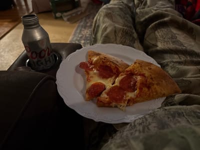 A casual scene shows two slices of pepperoni pizza on a white plate, placed near a can of Coors Light in a cup holder of a dark brown couch. The viewer sees legs covered with a patterned blanket. The focus is on the food and drink, indicating a relaxed and informal setting.