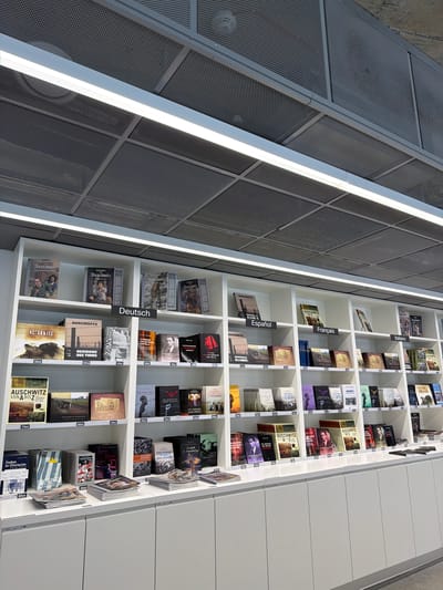 This is a high-angle, wide shot of a wall of bookshelves filled with books, primarily about Auschwitz. The bookshelves are white and neatly organized, with many books displayed vertically and some horizontally. The books appear to be in various languages, indicated by labels such as "Deutsch," "Español," "Français," and "Italiano." The cover art on many of the books features historical photographs or illustrations related to the Auschwitz concentration camp, with titles like "Auschwitz," "Residenz des Todes," and "Zurück in Auschwitz." Some books also have prices visible, indicated by "zł" (Polish Złoty).
To the left of the bookshelves, a textured, grey metallic wall with a grid pattern is visible, running parallel to the bookshelves. Two long, bright white fluorescent lights are mounted vertically along the edge of this metallic wall, illuminating the scene. The overall impression is of a library or museum exhibit dedicated to the history of Auschwitz, likely located in or near Oświęcim, Poland, as suggested by the presence of the Polish currency and titles referencing the location. The lighting creates a clean and somewhat sterile atmosphere, emphasizing the educational and somber nature of the displayed material.