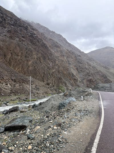 The image depicts a dramatic mountain pass landscape near Sumdo, India, under a cloudy sky. A paved road with a white dividing line curves to the right. To the left of the road, a steep, rocky mountainside rises sharply, showing layers of brown and gray rock formations. A river flows through a rocky ravine at the base of the mountain. Several telephone poles with wires are visible along the mountainside. The ground beside the road is covered in gravel and scattered rocks. A white post with the number "4" marked on it stands near the edge of the road. In the middle ground, a person in white clothing is standing near a guardrail. The overall atmosphere is rugged and remote, with signs of human infrastructure juxtaposed against the raw natural environment.