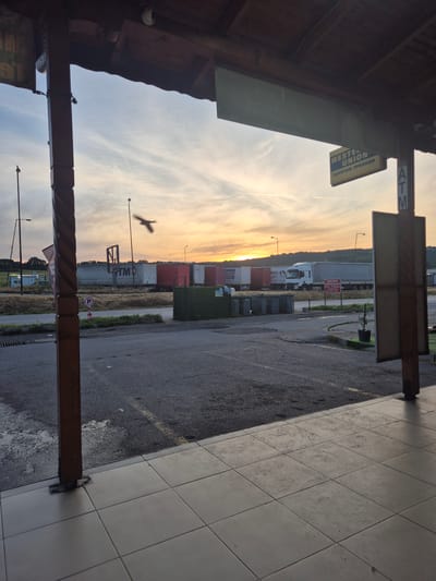 The image shows an outdoor scene in Lesovo, Bulgaria, at what appears to be a roadside stop or small commercial area at dusk or dawn. In the distance are shipping containers, a semi-trailer truck, and a sign for the Sivak Restaurant. Foreground elements include a partially visible building facade with tiled flooring and wooden support beams. There is signage for an ATM and Western Union. A bird is silhouetted against the sky. The ground is asphalt and slightly wet.