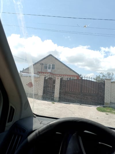 The image, taken from inside a vehicle, shows a residential scene in Rayevskaya, Russia. The steering wheel and part of the dashboard are visible in the foreground, indicating the view is from the driver's seat. Outside, a brick house with a triangular roof and white trim around the windows is seen behind a brick wall and ornate metal gate. Power lines stretch across the blue sky with scattered white clouds. The road in front of the gate is paved, showing signs of wear and dirt. The overall impression is one of a quiet residential area on a sunny day.
