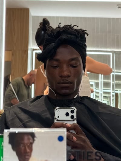 A young Black man with dreadlocks wrapped in a black headscarf is looking down at his phone. He is wearing a black cape. He has earbuds in. In the background, a woman is styling another person's hair. The location appears to be a salon. The image has a reflection of the man and the salon's interior.