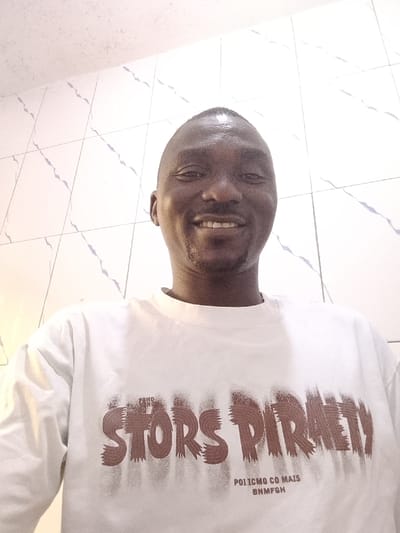 An adult male subject is depicted in a close-up, head-on shot in an indoor setting located in Akpajo, Nigeria. The subject is dark-skinned with a short, trimmed haircut, and a visible goatee or stubble on his chin. He is smiling directly at the camera, revealing his upper and lower teeth. He wears a white, short-sleeved t-shirt with distressed brown lettering on the front, which includes "STORS PIRATY" as a prominent element, and smaller, partially legible text above and below. The background consists of light-colored, possibly white, ceramic tiles arranged in a grid pattern on a wall. These tiles feature irregular, faint blue-grey vein-like patterns. A plain, light-colored ceiling is visible at the top of the frame. The lighting is bright and uniform.