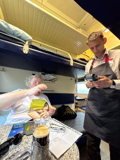 The image depicts two individuals inside what appears to be a train compartment. On the left, a person wearing a white shirt, a red lanyard, and a dark apron is holding a small black device. They have short, light-colored hair and are looking downwards. In the foreground, another individual with gray, curly hair and glasses is visible. They are wearing a white t-shirt with a graphic design featuring a woman's face. This person is holding a yellow object in their left hand and has their right hand raised, possibly in a gesture of greeting or explanation.
On a table in front of the second person, there are several items. A glass containing a dark liquid with a frothy top, likely a beverage, is present. Next to it is a silver foil-wrapped object and a clear plastic binder containing some papers, one of which has visible text. A smartphone is also on the table, displaying what appears to be an application or interface with colored graphics. The setting is dimly lit, suggesting it might be evening or the interior lighting is low. The walls of the compartment are lined with pale, horizontally slatted material, and there are dark blue upholstered seats. The overall impression is of a casual encounter or service interaction within a train.
