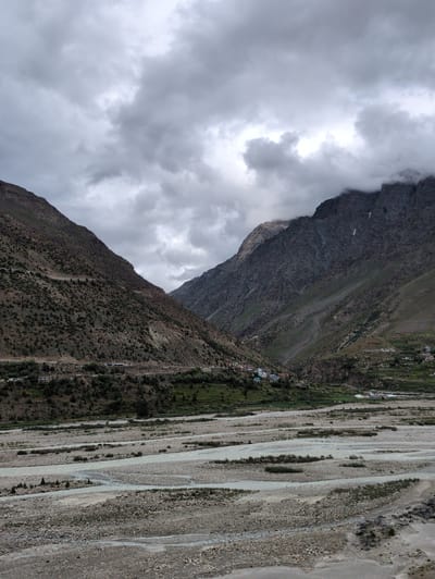 The image shows a wide, rocky riverbed in a mountainous valley. The river itself appears to be shallow and spread out, with numerous gravel bars and islands. The valley walls are steep and covered in sparse vegetation, with some evidence of human settlement on the lower slopes. In the foreground, the riverbed is dry and rocky. In the midground, the river flows in a braided channel. In the background, the valley narrows and the mountains rise steeply on either side, with a village visible on the left bank. The sky is overcast with heavy clouds, suggesting a moody or potentially rainy day. This scene is consistent with the landscape of Darcha, India, a village located in the Lahaul and Spiti district of Himachal Pradesh, known for its high-altitude valleys and glacial rivers.