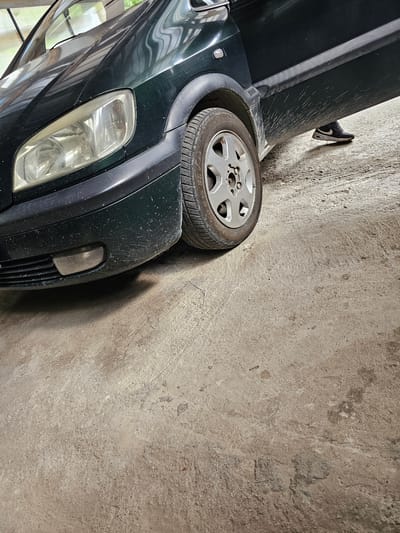 A dark green vehicle is parked at an angle next to a concrete wall in Erma Reka, Bulgaria. The front passenger side of the car, including the headlight, bumper, and a partially obscured wheel, is visible. A shoe is positioned on top of the car near the wall. The setting appears to be an enclosed or semi-enclosed space, potentially a garage or alleyway. The car and the concrete wall are dusty.
