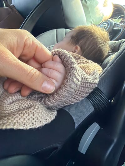 In Karacha-Elga, Russia, the image depicts a close-up scene inside a vehicle. An adult's light-skinned right hand, with visible fingernails, is holding the left hand of an infant. The infant's small, light-skinned fingers are curled around the adult's index finger. The infant, displaying light brown hair on its head, is partially visible and appears to be resting in a dark-colored car seat or infant carrier. A light brown, knitted blanket with a distinct patterned weave covers the infant's body, extending to the area of the hands' interaction. The car seat features dark gray and black upholstery, a visible black strap, and a light gray plastic release mechanism on its side. The background shows blurred elements of the car's interior, including a steering wheel and parts of the dashboard, under natural light conditions.
