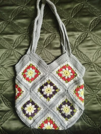 A handmade crocheted tote bag with a granny square pattern is displayed against a quilted green fabric background. The bag is made of light gray yarn, with each square featuring a floral motif in various colors, including red, yellow, white, purple, and green. Two straps extend from the top of the bag. The background fabric has a repeating circular pattern created by stitching. The image is a top-down view, and the lighting suggests it was taken indoors or in a shaded outdoor area. The specific location in Калодзішчанскі сельскі Савет, Belarus, cannot be determined from the visual information provided in the image. There are no people or actions depicted.