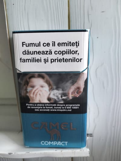 A pack of Camel cigarettes sits on a white, textured surface, possibly a shelf. The pack is primarily teal with a white panel on the front. The white panel features text in black and a photograph. The text reads, "Fumul ce îl emiteți dăunează copiilor, familiei și prietenilor." Below this text is a photograph of a child covering their mouth and nose with their hands, appearing distressed, with smoke around them. To the right of the child, a hand is seen holding a lit cigarette. Below the photograph, there is smaller text in white stating, "Pentru a obține informații despre programele de renunțare la fumat, sunați la 0 800 10001 sau accesați www.impsd.md." The bottom teal section of the pack displays the Camel logo, which is the brand name in large, brown letters above a silhouette of a camel. Beneath the Camel logo, the word "COMPACT" is written in white. The image is lit from the front, casting no significant shadows.