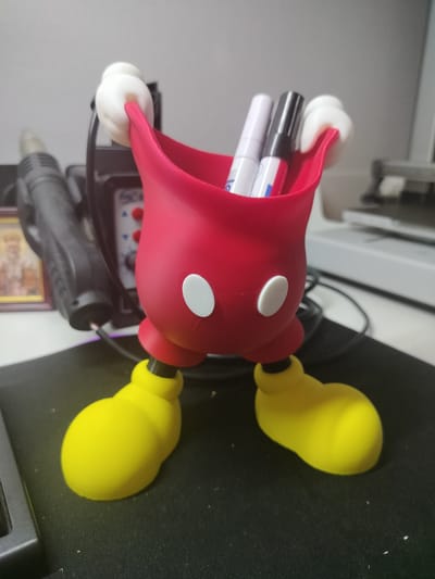 A 3D-printed holder shaped like Mickey Mouse's torso and legs stands on a black mousepad. The red torso has two white circles resembling eyes and holds two markers, one black and one white. The character's yellow feet are positioned apart. In the background, to the left, is a heat gun with a black handle, and to the right, a portion of a white electronic device is visible. The scene is indoors, on a desk, with no specific indication of Mokhsogollokh, Russia.