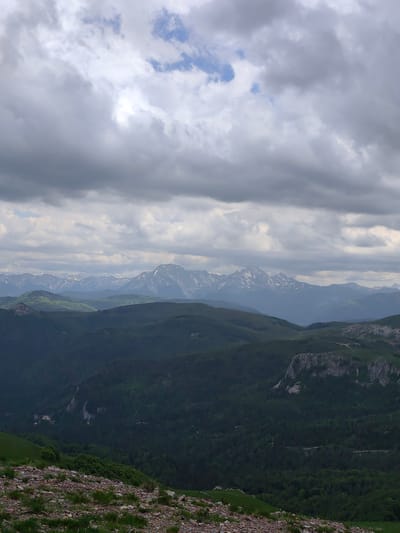 The image presents a panoramic mountainous landscape in Sedlačka gora, Montenegro, under a predominantly overcast sky. The sky occupies the upper approximately one-third of the frame, filled with dense, textured grey clouds, with small, fragmented patches of lighter blue visible.
The mid-ground features multiple layers of rolling hills and valleys. The nearest hills are covered in a dark, dense forest of evergreen trees. Within this forested area, several grey rocky cliffs and outcrops are visible, particularly on the right side. Faint light-colored lines, consistent with unpaved roads or tracks, snake through the forested terrain. Above these immediate hills, layers of lighter green, possibly grasslands or less densely forested slopes, recede into the distance.
The background is dominated by a long, rugged mountain range stretching across the horizon. These distant peaks are distinctly snow-capped, with white snow visible on their upper slopes against a dark, rocky mass. Atmospheric perspective renders these mountains in muted blue-grey tones.
The immediate foreground, covering the bottom approximately 15-20% of the image, consists of uneven, rocky ground with scattered small stones and sparse clumps of green grass. No human figures, animals, or significant man-made structures are present within the scene. The lighting is diffused, indicating an overcast or heavily cloudy day, with soft shadows and consistent ambient light across the landscape.