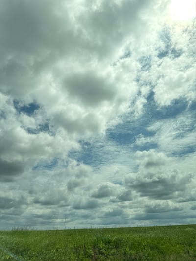 The image depicts a vast, cloudy sky over a green, grassy field. The clouds are varied, with some appearing bright and white against patches of blue sky, while others are darker and more ominous. The sun is visible, though partially obscured by clouds, creating a bright glow in the upper right portion of the frame. The field in the foreground is covered in tall, green grass, suggesting an open, rural environment. There are no people or man-made structures visible in the scene. The image appears to be a landscape photograph taken from a low vantage point, looking up towards the sky. There is no information available to confirm the location in the city of Maksimovo, Russia.