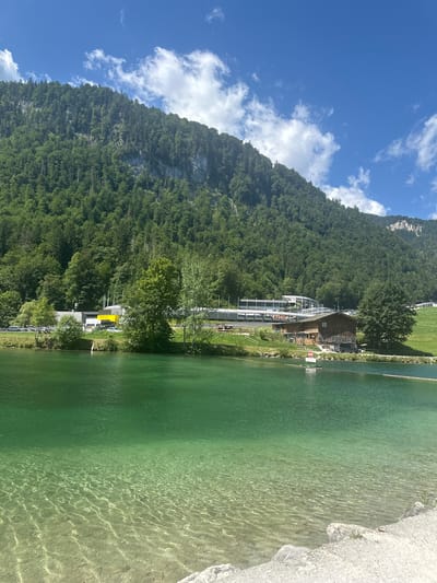 This is an outdoor, eye-level, vertical shot of a clear, turquoise lake in Schönau am Königssee, Germany. In the foreground, the lake bottom is visible through the shallow, clear water, with sandy and rocky textures. The water transitions to a deeper, opaque green in the mid-ground. Along the far shore of the lake, a grassy bank is lined with trees. Behind the trees, there is a wooden building with a sloped roof and a modern, white building with a curved roof, possibly a visitor center or administrative building. Further back, a steep, densely forested mountain rises, its peak partially obscured by white, puffy clouds against a bright blue sky. A road with parked cars is visible between the lake and the buildings.