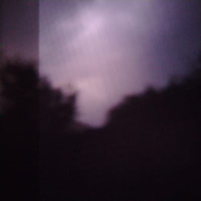 The image is a dark, abstract photograph that appears to be taken at night or in very low light conditions. The dominant colors are deep purples and blacks, with a faint, hazy area of lighter purple or white in the upper center of the frame. There are blurred, dark shapes at the bottom and on the left and right sides that suggest the presence of foliage or structures, but they are too indistinct to identify clearly. The overall impression is one of obscurity and a lack of detail, making it impossible to discern any specific subjects, actions, or interactions. The image does not provide any visual cues that would allow for its location to be identified in the city of Kuyoshli (Solnechny), Uzbekistan.