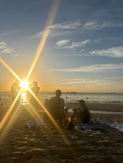 The image depicts a coastal beach scene at sunset in Black Rock, Australia. The sun, low on the horizon to the left-center, generates significant lens flare across the frame, silhouetting figures and casting a golden hue over the environment. The sky is a gradient from golden-orange at the horizon to light blue overhead, with scattered wispy white clouds. The calm ocean surface reflects the sunlight, and multiple dark figures are visible wading or swimming in the water. In the distance on the right, block-like structures, likely coastal defense or pier remnants, are visible, along with a vertical marker pole and faint silhouettes of distant vessels.
On the sandy beach in the mid-ground, five individuals are present. Three figures are standing near the water's edge on the left, largely silhouetted by the sun. One appears to be raising an object to their mouth. To the right, two individuals are seated facing the ocean, with their backs to the camera. One is seated in a dark, foldable beach chair. The other is seated directly on the sand to their right, wearing a dark sleeveless top with a white number "7" on the back and a dark baseball cap. A yellow and black bag, a striped towel, and a white towel are positioned on the sand near the seated figures. The foreground features textured sand with visible footprints.