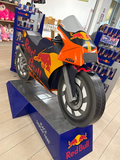 In the city of Due Carrare, Italy, a Red Bull branded motorcycle cutout is displayed on a blue pedestal. The cutout is orange and black, featuring the Red Bull logo and the number "1". Behind the motorcycle, shelves are stocked with Red Bull cans. The environment appears to be a retail store, with wooden flooring visible. No people are present in the immediate foreground of the image.