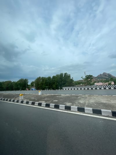 The image depicts a road in Addakal, India, under a cloudy sky. The road is asphalt with a white line marking its edge, bordered by a curb with alternating black and white markings. Further to the right, a divided highway features a chevron-patterned barrier. Trees line the road, and in the background, a rocky hill with buildings is visible. Two concrete posts, one yellow and one white, stand near the roadside, with the yellow post bearing the number "118". The scene appears to be an outdoor, daytime view.