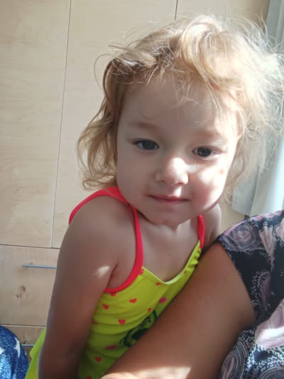 This media file shows a close-up, waist-up shot of a young Caucasian child with fair, tousled hair. The child is looking towards the camera with a slight smile and appears to be held by an adult. The child is wearing a bright yellow swimsuit with small red hearts. The background consists of a wooden closet with metal handles. Sunlight is streaming in from the right, casting shadows on the child's face and arm. A portion of an adult's patterned clothing is visible on the right side of the frame. The scene appears to be indoors, possibly in a bedroom, given the closet. The lighting suggests it is daytime. There is no visible text in the image.