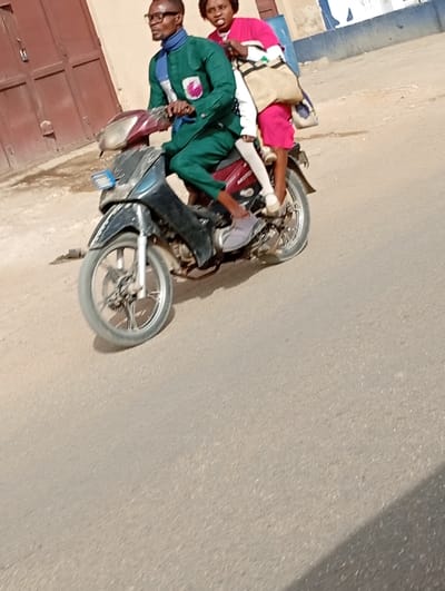 An image captured in Gayawa, Nigeria, depicts two individuals riding a single motorcycle on a paved road under daylight conditions. The motorcycle, dark-colored with a maroon rear section and visible "MOTO" branding, is in motion, evidenced by the slight blur of its front wheel.

The driver is a male individual, appearing to be in his 20s or 30s, wearing eyeglasses, a dark green long-sleeved tunic with a light-colored design on the chest, a blue and white patterned scarf or neck wrap, and dark green trousers. He has light-colored footwear on his right foot and appears barefoot on his left foot, which is positioned on the motorcycle's footrest. His gaze is directed forward and slightly to the left.

Seated behind the driver is a female individual, also appearing to be in her 20s or 30s. She wears a bright pink long-sleeved top with a white stripe pattern on the sleeves, bright pink trousers or capris, and light-colored closed-toe shoes. She carries a light-colored shoulder bag with dark straps, positioned over her right shoulder. Her expression is neutral, and she looks towards the right side of the frame. Her legs are positioned over the motorcycle's rear seat, with her feet on the passenger footrests.

The background shows a light brown or tan building with a closed brown metal door on the left. Further to the right, portions of a light-colored wall with blue markings are visible. The road surface is light grey asphalt. Shadows are cast from the left side, indicating the sun's position.