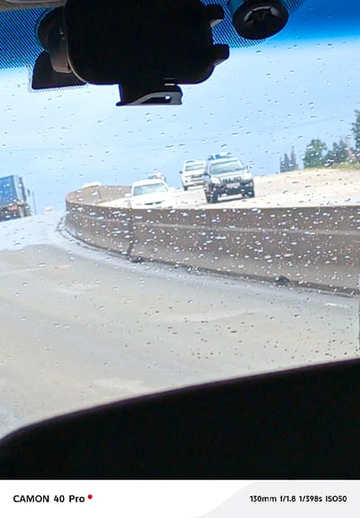 The image, captured from the interior of a vehicle looking outward through a rain-splattered windshield, depicts a multi-lane highway in Sigona ward, Kenya. Visible through the windshield's upper edge are a dark overhead mounting bracket and a circular device. The road surface appears wet. On the elevated roadway, traffic proceeds along a rightward curve. From foreground to background, and right to left, a dark-colored SUV with a roof-mounted fixture, followed by a white SUV, and then a white sedan are observed. Further left in the distance, a blue semi-truck trailer is partially visible. A concrete median barrier separates the main thoroughfare from a lower, unseen road segment or shoulder. The horizon shows a bright, overcast sky and distant green foliage. No individuals are discernible.