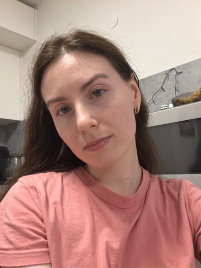 An individual is depicted indoors within what appears to be a kitchen setting in the city of Ковалёво, Russia. The subject is a female, seen from the mid-chest upwards, looking directly at the camera with a neutral expression. She has long, dark brown hair, fair skin, light-colored eyes, and is wearing a plain, coral-pink short-sleeved t-shirt. A small, gold-colored hoop earring is visible in her right ear. In the background, a white wall or ceiling is visible above grey tiled backsplash. A string of small, white LED fairy lights is affixed to the tiled wall. Below the lights, a light-colored appliance, possibly a microwave, is present, with a translucent, fluted bowl containing indeterminate contents resting on top of it. White overhead cabinets are partially visible in the upper left. Stacked dark-colored objects are partially visible in the far-left background.