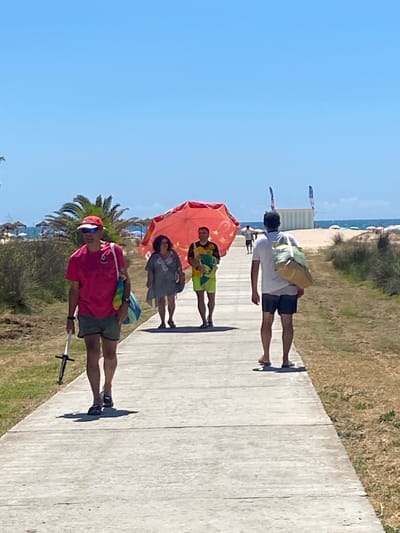 The image depicts a sunny day at a beach in Conil de la Frontera, Spain. A concrete pathway runs through the scene, bordered by dry grass and some vegetation. Four individuals are walking along the pathway. In the foreground, a person wearing a bright pink t-shirt, a red cap, and sunglasses, and dark shorts, is walking away from the camera, holding a long object with a black handle. They are carrying a colorful striped bag over their shoulder. Behind this person, a woman in a light gray, loosely fitting dress is walking, holding a large, open coral-colored umbrella. To her right, a man in a yellow and black patterned shirt and lime green shorts is walking, carrying a folded towel. Further down the path, a man in a white t-shirt and dark shorts is walking away from the camera, carrying a large, tan and green duffel bag over his shoulder. In the background, the beach is visible with numerous umbrellas and lounge chairs, and the blue sea under a clear sky. Several flags are planted in the sand, some appearing to be for water sports or beach activities. The overall impression is of a warm, leisurely day at the coast.