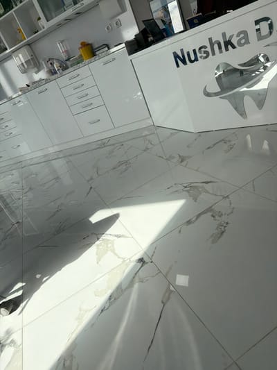 This image depicts the interior of a modern dental clinic, identified by signage as "Nushka D," located in the city of Hvostiane, Bulgaria. On the left, a long, white, high-gloss cabinetry system with multiple drawers featuring chrome pull handles extends along a wall. A white countertop above the drawers holds a clear plastic water dispenser, a yellow lidded biohazard waste bin, and various smaller containers. Open shelving is visible above the countertop. To the right, a white reception desk displays the name "Nushka D" in reflective metallic lettering, accompanied by a reflective metallic tooth-shaped logo. A laptop and a computer monitor are positioned on the desk. The floor is covered with large, polished white ceramic tiles featuring distinct gray and black marble-like veining. Direct sunlight streams into the space, casting sharp, angular shadows across the reflective floor. No individuals are visible in the scene.