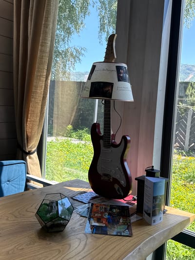 The image captures an indoor setting in Ozernoe, Russia, featuring a wooden table next to a large window with a view of lush green trees and a bright blue sky, indicating a sunny day.

The primary objects on the table include a red electric guitar with a white pickguard and "Terris" visible on its headstock. A piece of paper or fabric adorned with small images and Cyrillic text is draped over the guitar. Partial legible text includes "Я часто вижу прих В смотрению на деле" (I often see... in action/practice) and "Хонхилон Хай! Чегкость дока" (Honkhilon Hai! Dock clarity).

Further along the table, a black stand holds a brochure displaying a scenic mountain landscape and QR codes, suggesting information about the local area or amenities. Nearby, several other printed cards or postcards are visible, one prominently featuring the Cyrillic text "БАННЫЙ СПА БУДУЩЕГО" (BATH SPA OF THE FUTURE). A geometric glass terrarium containing small plants adds a decorative touch to the wooden surface.

A comfortable-looking blue upholstered chair with a button-tufted back is partially visible beneath the table. A light-colored, pleated curtain is tied back, allowing the bright natural light to stream in. The scene appears to be a cozy corner designed for relaxation, possibly in a guesthouse or a nature-oriented retreat, blending indoor comfort with outdoor scenery. No specific activity is depicted, giving the impression of a peaceful, ready-to-use space.