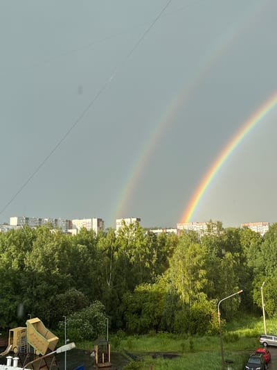 This image captures a double rainbow arching across a pale, overcast sky, viewed from a slightly elevated position. The scene is dominated by the vibrant colors of the rainbow on the left and a dense canopy of lush green trees on the right. Beyond the trees, a series of apartment buildings are visible, their facades a mix of white, grey, and muted colors.
In the foreground and along the right edge of the image, the vibrant green foliage of trees creates a natural border. Interspersed within the trees and to the right are signs of urban infrastructure, including streetlights and a playground structure. A few cars are parked along a path or road to the far right, including a red car and a dark sedan. Thin, dark lines of what appear to be wires or cables crisscross the sky in the foreground.
The weather appears to be overcast, with the sky being a uniform light grey, which is typical of conditions that produce rainbows after rain. The presence of the rainbow itself suggests recent or ongoing precipitation. The overall mood is one of natural beauty contrasted with a typical urban or suburban environment. The time of day is difficult to determine precisely due to the overcast conditions, but the lighting suggests it is daytime. The location is identified as Bratsk, Russia. No visible text is present in the image.