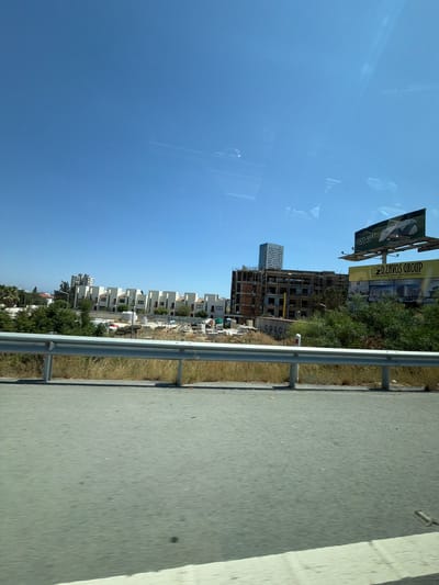 This is a landscape-oriented photograph taken from a moving vehicle, likely a car, as indicated by the road and barrier in the foreground and the slight motion blur. The scene depicts a construction site with several buildings under various stages of development. In the background, there are two prominent billboards. The left billboard displays "COCO-MAT sleep on natur" with an image of a mattress. The right billboard advertises "DZAVOS GROUP LAND & BUILDING DEVELOPERS SINCE 1980," featuring images of modern residential properties.
Several rows of townhouses, mostly white with dark-framed windows, are visible, suggesting a residential development. One taller, modern building with a white facade and numerous windows stands out amongst them. The overall setting appears to be a sunny day, given the clear blue sky and bright lighting. The presence of construction materials and scaffolding on the partially built structures indicates ongoing development activity. The view suggests a location on the outskirts of a developed area, possibly near a highway or main road, given the presence of the road and guardrail. The Mouttagiaka Community in Cyprus is the identified location context.