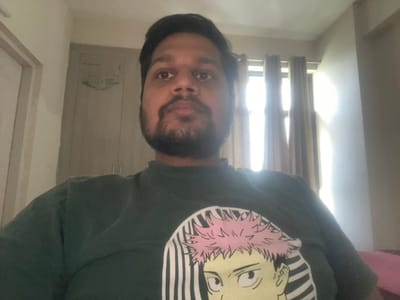 The image shows a young man with a beard, wearing a green t-shirt with a cartoon character graphic, sitting in a room in Greater Noida, India (latitude: 28.476, longitude: 77.546). The room has white walls, a white ceiling, and a closed door to the left of the frame. There is a large window on the right side of the frame, with curtains drawn. The room is brightly lit by natural light from the window.  The man is looking directly at the camera.  His expression is neutral, his gaze is steady, and his shoulders are relaxed. He is sitting alone in the room.  The image is captured from a slightly elevated angle, and the man is positioned in the center of the frame.
