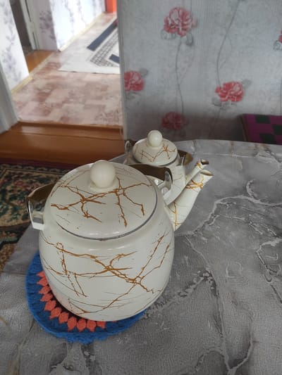 The image shows a cream-colored teapot with gold marbling, sitting on a crocheted blue and orange coaster on a table. A second, smaller teapot is visible behind it. The table is covered with a grey, textured tablecloth. In the background, a wall with a floral wallpaper pattern featuring pink roses is visible. To the left, a doorway leads to a hallway with wooden flooring and a patterned rug. The setting appears to be a domestic interior, likely a home in Некрасовка, Kazakhstan. There are no people or animals present in the image.