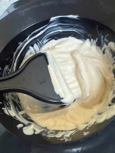 This media file shows a close-up overhead view of a black bowl filled with a creamy, light-colored substance, likely hair dye or a similar cosmetic mixture. A black plastic applicator brush is submerged in the mixture, with some of the substance clinging to its bristles and handle. The bowl's interior shows streaks of the lighter substance against the dark background, indicating it has been mixed or stirred. The texture of the mixture appears smooth and slightly viscous. There is no visible text in the image. The lighting suggests an indoor setting, likely during the daytime. The specific location context of Hoofddorp, Netherlands, is provided but not visually evident in the image itself. The scene focuses solely on the preparation of a cosmetic product.