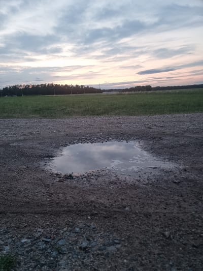 The image shows a puddle of water on a gravel path in Pohránov, Czechia, during a cloudy sunset. The foreground features the textured gravel and the reflective puddle. Beyond the path, a green field stretches towards a line of trees and distant structures, possibly including power lines, under a sky filled with dramatic, streaked clouds tinged with the soft colors of twilight. The scene is devoid of any people or animals.