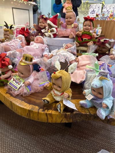 A detailed view captures a retail display in Santa Cruz Aquiahuac, Mexico, featuring numerous baby dolls arranged on a thick, polished wooden slab table. Approximately twenty to thirty dolls are visible, varying in skin tone and dressed in diverse costumes, predominantly with Christmas or festive themes. Outfits include Santa suits, reindeer costumes, elf attire, unicorn onesies, tutus, and holiday bows. Some dolls are positioned directly on the table, while others sit atop white cardboard boxes. Several dolls are individually wrapped in clear plastic packaging with decorative ribbons, indicating they are merchandise for sale. White price or brand tags, including one clearly visible for "Furever Dreaming Kids Entertainment," are attached to multiple dolls. The blurred background suggests an indoor commercial setting, possibly a market stall or store, with additional merchandise and decorative string lights visible in the upper frame. A person's blurred figure is barely discernible in the upper left. The floor beneath the table is carpeted in a neutral tone.