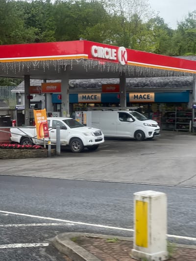 The image depicts a Circle K gas station in an unknown location in Ireland. The station features a prominent red roof with white and orange trim, adorned with decorative string lights. Underneath the canopy, several gas pumps are visible. A white pickup truck is parked to the left, displaying an orange sign that reads "Circle K extra sign up and save." To the right of the pickup truck, a white van is parked. The background shows trees and buildings, including signage for "MACE" and "miles." The scene appears to be during daylight hours.