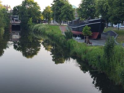The image shows a still body of water reflecting trees and buildings. On the right bank, a black ship with red hull plating is displayed on land. The ship has the name "HERMINE" written on its side. The surrounding area is green with trees and grass. In the background, there are buildings and a bridge. The setting appears to be a public space in Cuxhaven, Germany, possibly a waterfront park or museum.