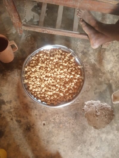 An overhead view captures an indoor scene in Gudige Sabo, Nigeria. A circular, silver-colored metal tray, approximately 20 inches in diameter, is positioned centrally, filled with a dense layer of small, irregularly shaped, light brown pieces resembling fried dough or snacks. To the upper left of the tray, a light brown, handled mug rests on the floor. In the upper right quadrant, a pair of bare human feet are visible, partially obscured by the dark brown, horizontal and vertical bars of a wooden or metal furniture frame. To the lower right of the tray, an irregular mound of light brown, granular material, consistent with dirt or sand, sits on the floor. The floor itself is a mottled grey and light brown concrete or cement surface, exhibiting various textures and areas of wear. No direct action is depicted beyond the placement of these items and the resting feet.