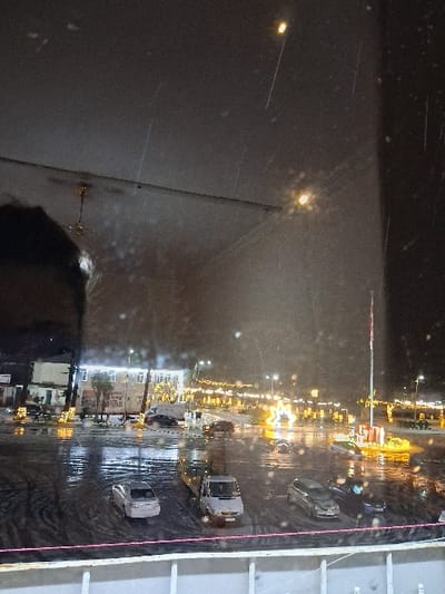 The image presents a nighttime outdoor scene in Lanchkhuti, Georgia, characterized by falling snow or sleet and wet surfaces reflecting artificial light. The perspective is from an elevated interior, looking through a window, evident from the white railing at the bottom, reflections of an internal light fixture, and the dark silhouette of a person's head on the upper left.
The ground consists of a large, wet asphalt or paved area, likely a public square or parking lot, marked with numerous tire tracks. Several vehicles are visible: a silver sedan parked on the left, a white commercial van in the center, and a dark sedan on the right. Additional vehicles are partially obscured further back.
In the background, buildings with illuminated windows and facades line the street. The area is extensively lit by streetlights and decorative yellow-orange string lights draped on trees and other structures. A prominent star-shaped light installation is visible towards the right-center. A tall flagpole with a red and white striped base and a partially visible flag stands near this area. No individuals are clearly discernible.