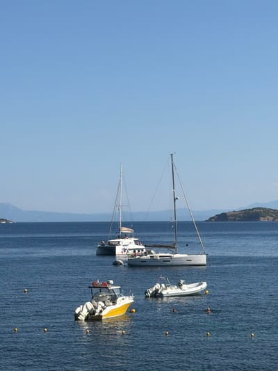This is a vertical, wide-angle shot of a clear blue sky over a body of dark blue water, with a mountainous coastline in the distance. Several boats are docked or anchored in the water. In the foreground, a white catamaran is moored, its mast and rigging clearly visible. To the right of the catamaran, a rigid inflatable boat (RIB) is tied alongside. Further to the right and slightly behind, a yellow and white motorboat is anchored. Scattered yellow buoys are visible in the water around the boats. The sun is high in the sky, indicating it is likely midday. The water appears calm, and the sky is cloudless, suggesting fair weather. The scene evokes a sense of peaceful summer boating. This image was captured in Skiathos, Greece. No visible text is present.