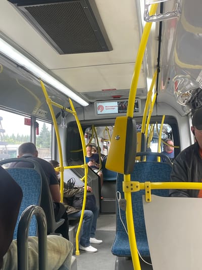 The image shows the interior of a bus in Yakovlevskoye, Russia. The scene is viewed from the perspective of a seated passenger, looking towards the front of the bus. Several people are visible, seated and standing. The bus has blue patterned seats and bright yellow handrails, characteristic of public transportation.
One passenger in the foreground is facing away from the camera, their head and shoulders visible. Further back, a man in a blue shirt and jeans is seated, his legs extended. Another person, possibly also a man, is leaning his head against the window. In the distance, other passengers are partially visible.
A screen on the bus displays text in Cyrillic, which appears to be an advertisement or information related to "Московский транспорт" (Moscow Transport). The lighting suggests it is daytime, and the view through the windows shows trees and a fence, indicating an outdoor environment. The overall atmosphere seems ordinary, depicting a typical bus journey. There are no overt signs of any particular event or strong emotions. Objects like a black vent on the wall and various poles and fixtures of the bus are also visible.
