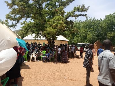 The image shows a gathering of people in an open-air space in Zum, Nigeria. The coordinates provided place the location in the rural outskirts of the city, likely a village or town. There are at least 30 people present, mostly women and children, seated in rows on green plastic chairs. There is a large white tent in the foreground, and a smaller blue tent behind the crowd. A large green tree provides shade in the center of the image. Behind the crowd, there is a building with a white roof and a white wall, indicating a possible public or community building. A white bus is parked further back, to the right. Several people are standing, interacting with each other, possibly discussing something or greeting each other. The ground is dusty and dry, and the environment is typical of a rural setting in Nigeria.