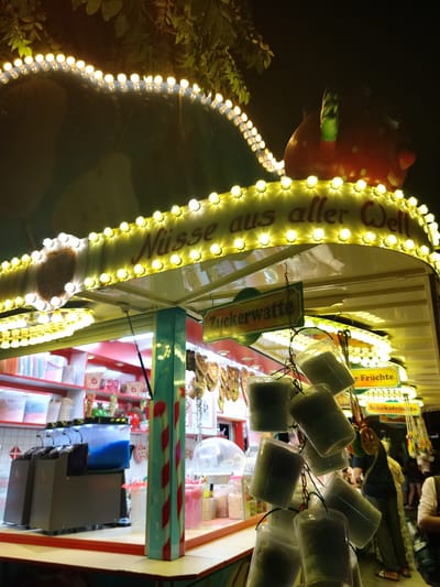 This is a nighttime, eye-level, medium shot of a brightly lit food stall at a festival or market in Cottbus - Chóśebuz, Germany. The stall is adorned with strings of yellow, incandescent lights that illuminate the signage and the structure. A large, cartoonish red strawberry figure with green leaves is mounted at the top right of the stall. Above the main counter, a yellow sign with red lettering reads "Nüsse aus aller Welt," translating to "Nuts from all over the world." Below this, a green and yellow sign reads "Zuckerwatte," which means "cotton candy." Several clear plastic bags filled with what appears to be cotton candy are hanging from chains. In the background, shelves are stocked with various food items and containers. To the right, a person wearing a dark shirt and light pants is visible, with other festival-goers in the distance. The stall's counter is a light wood color with a red trim.