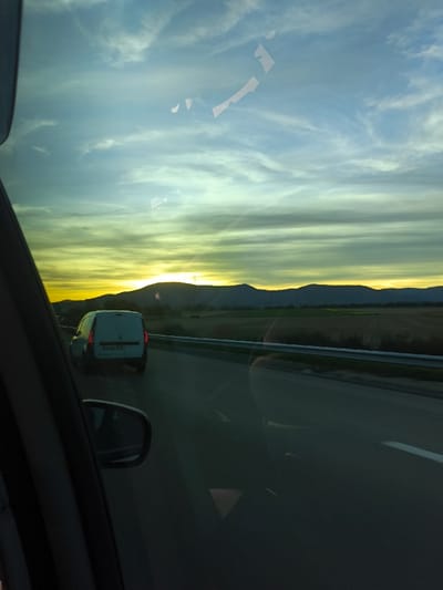 The scene captures an exterior view from within a vehicle, observed through the front passenger-side window, on a multi-lane paved road in the vicinity of Niedernai, France. The time of day is either sunrise or sunset, characterized by a low sun casting an intense yellow-orange glow on the horizon, silhouetting a range of dark, undulating hills. The sky above features horizontal cloud streaks, transitioning from golden hues near the horizon to light blue and grey. To the right of the road are expansive agricultural fields, separated by a metal guardrail. A white cargo van, displaying a French license plate (partial registration visible: 'DA-XXX-XX'), travels in the same direction in the lane ahead. Reflections from the interior are present on the window, and a portion of the vehicle's side mirror is visible in the lower left quadrant. No individuals are depicted.