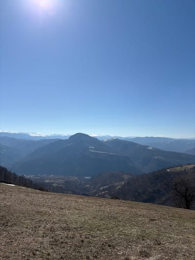The image displays a panoramic mountainous landscape with a clear, bright blue sky occupying the upper two-thirds of the frame, featuring a visible sun flare in the top-left corner. The foreground presents a gently sloping, dry grassy field with patches of brown and light green vegetation, along with minimal isolated snow patches on the far left. Below the foreground, a valley descends, revealing a settlement identified as Babintsi, Bulgaria, consisting of scattered residential and commercial structures amidst forested slopes. Utility poles are faintly discernible within the valley. Multiple layers of mountains extend into the background; midground mountains are primarily covered in bare deciduous trees, while the most distant peaks, arrayed along the horizon, exhibit prominent snow caps. No human figures or active subjects are present in the scene.