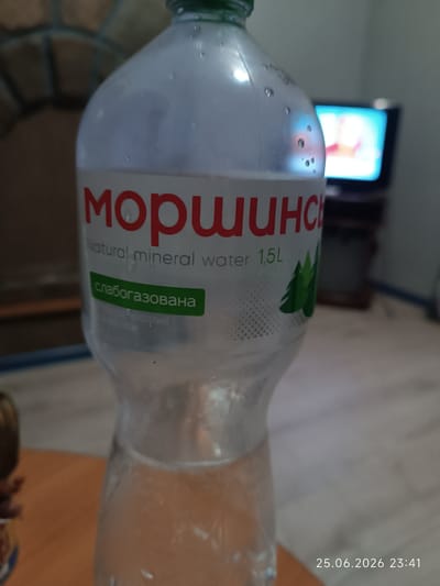 A clear plastic bottle of "Моршинска" natural mineral water, 1.5L, is prominently displayed in the foreground. The label is white with red lettering for the brand name, and white lettering for "Natural mineral water 1.5L". A green banner with Ukrainian text "слабогазована" (low-carbonated) is present below the main text. The bottle is partially filled with water and shows condensation droplets on its surface. In the blurred background, a television screen displays a colorful image, and a stone fireplace is visible to the left. The timestamp "25.06.2026 23:41" is in the lower right corner of the image. The scene appears to be indoors, possibly in a domestic setting. There are no people visible in the image. The location is specified as Bolhrad, Ukraine.