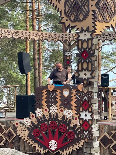 This image depicts an outdoor DJ performance at an event in Luzhki, Russia, characterized by a rustic and nature-inspired aesthetic. In the foreground, a DJ, a man with glasses and a beard, is actively mixing music at a console. He is wearing a dark t-shirt. Beside him, a young woman with light brown hair stands looking towards the DJ. They are positioned behind a DJ booth decorated with intricate, laser-cut wooden panels featuring geometric patterns in brown, red, and white. Two large black speakers are visible, one on a stand to the left of the DJ and another smaller speaker on a table to the right. The background is dominated by tall pine trees, suggesting a forest or park setting. Further decor includes more laser-cut wooden elements with symbolic patterns, reminiscent of traditional folk art, suspended above and integrated into the stage structure. The overall scene conveys a festive, open-air atmosphere.