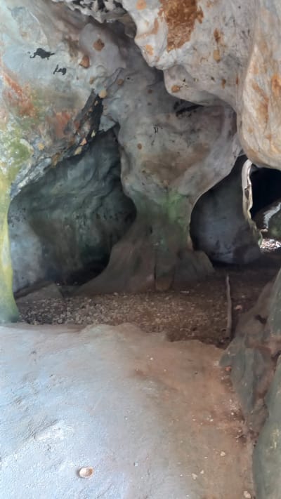 The image depicts the interior of a cave or rock formation, possibly located near Thap Put, Thailand. The walls are made of gray and brown rock, with irregular shapes and textures. A vertical column composed of pebbles or small rocks is visible on the right side of the frame. The lighting is dim, with shadows obscuring some areas of the cave. No people or animals are visible.