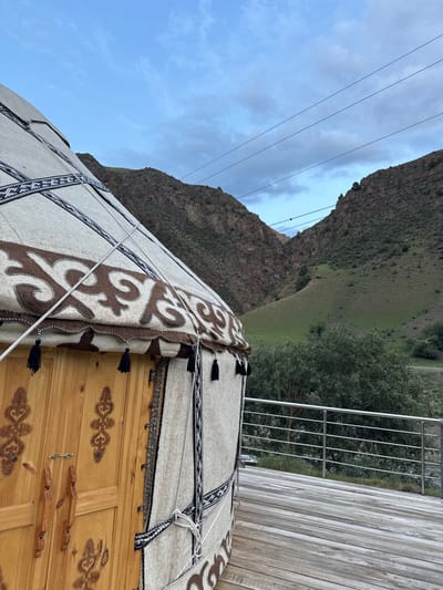 The image captures a close-up view of a traditional yurt, its intricately patterned felt exterior and wooden door prominently featured in the foreground. The yurt is situated on a wooden deck, complete with a modern metal railing, suggesting a tourist accommodation or hospitality setting. Beyond the yurt, a lush, green mountainous landscape unfolds under a cloudy sky, hinting at the natural beauty of the Kyzyl-Korgon region in Kyrgyzstan. The scene appears serene, with no people visible, and the overall impression is one of cultural immersion and natural tranquility.