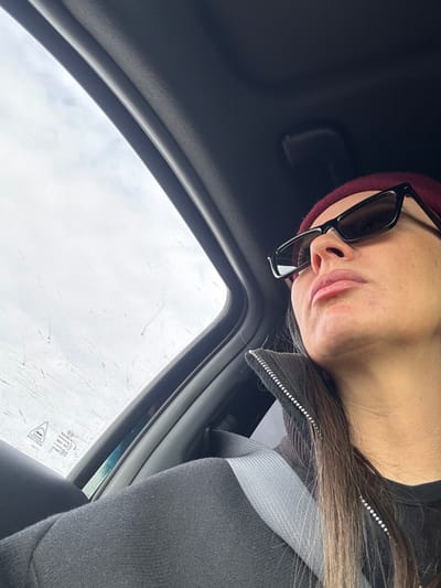 The image depicts a close-up, low-angle perspective from inside a vehicle, focusing on the right profile of an adult female. She is seated, looking upwards and to her left, out of a car window. Her head is adorned with a dark red beanie, and she wears dark, rectangular-framed sunglasses. Her lips appear full, and a defined jawline is visible. Strands of dark hair fall alongside her face and neck. She is dressed in a dark-colored garment, possibly a jacket or top, featuring a zippered collar with small, reflective elements on the zipper line. A light gray car seatbelt is visible across her right shoulder and chest. The vehicle's interior features a dark grey or black headliner and a dark A-pillar. Through the car window, an overcast sky is visible, with numerous linear streaks and smudges on the glass, consistent with dirt or rain residue. The precise location within Yurkovo, Russia, is not ascertainable from the visual data provided in the image.